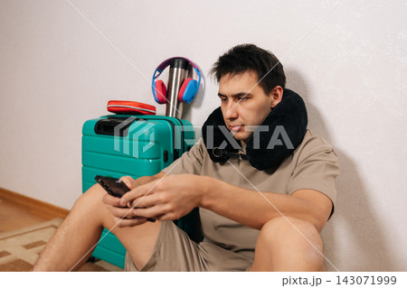 Male traveler sitting on floor with neck pillow and smartphone, having a delay, waiting for departure, feeling impatient while checking updates on mobile device during trip. 143071999