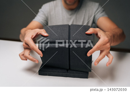 Close-up of man holding black acoustic foam panel, examining sound absorbing material for noise control, soundproofing, insulation in dark recording studio or home environment. Close-up of man holding black acoustic foam panel, examining sound absorbing material for noise control, soundproofing, insulation in dark recording studio or home environment. 143072030