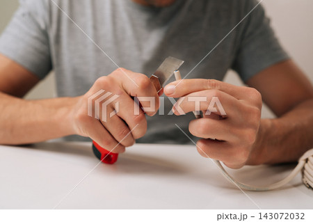 Close-up of electrician hands splitting communication cable with utility knife sitting at white table, preparing data wires for connection and installation, performing electrical repair work. Close-up of electrician hands splitting communication cable with utility knife sitting at white table, preparing data wires for connection and installation, performing electrical repair work. 143072032