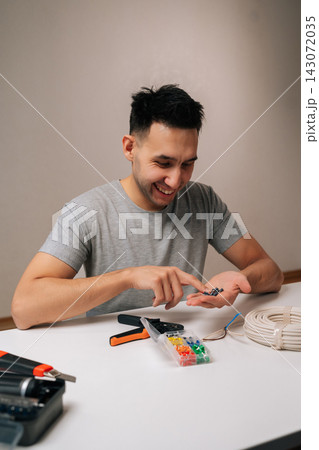 Cheerful electrician male selecting wire ferrules from a plastic organizer next to coiled electrical cable sitting at white table, depicting diy, home improvement and professional electrical work. Cheerful electrician male selecting wire ferrules from a plastic organizer next to coiled electrical cable sitting at white table, depicting diy, home improvement and professional electrical work. 143072035