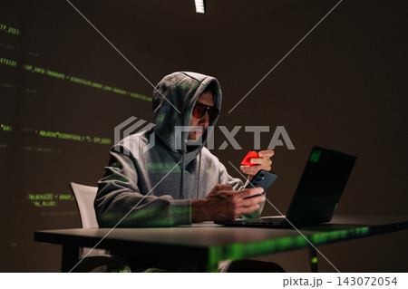 Dangerous hooded hacker male in sunglasses using smartphone and holding red credit card, surrounded by green code visuals suggesting cybercrime, fraud and identity theft. 143072054