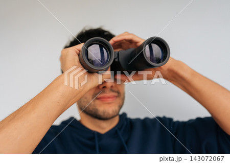 Close-up portrait of curious man holding binoculars to eyes, looking intently forward, actively seeking new perspectives, observing prospects, and exploring opportunities for future. Close-up portrait of curious man holding binoculars to eyes, looking intently forward, actively seeking new perspectives, observing prospects, and exploring opportunities for future. 143072067