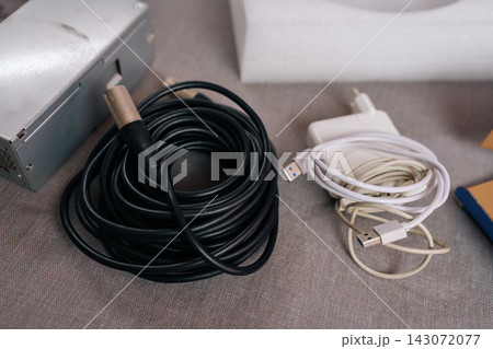 Close-up of wires and equipment. Coiled black XLR audio cable and white USB charger on gray fabric with power supply. Concept for technology, connectivity, sound engineering, electronic waste. Close-up of wires and equipment. Coiled black XLR audio cable and white USB charger on gray fabric with power supply. Concept for technology, connectivity, sound engineering, electronic waste. 143072077