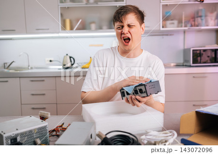 Man makes disgusted face cleaning dusty electronic part with blue cloth. He sneezes from dust allergy or frustrated with tech repair. Messy foreground with computer components in modern kitchen. 143072096