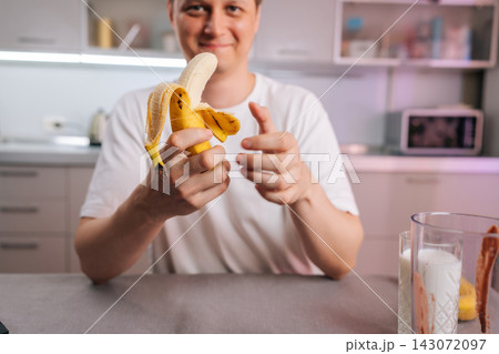 Happy man holding peeled banana prepares healthy breakfast smoothie in modern kitchen. Shallow depth of field focuses on fresh fruit. Concept for healthy lifestyle, diet, nutrition and wellness. 143072097