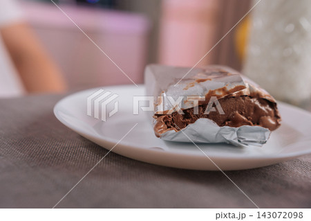 Chocolate chocolate coated ice cream bar unwrapped on white plate, silver foil peeled back to reveal creamy frozen center indulgent, rich dessert close-up studio shot. 143072098