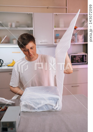 Focused young man in white t-shirt removes packaging from new electronic device. He assembling or repairing computer hardware at home, sitting at kitchen table. Concentrated DIY project. 143072099