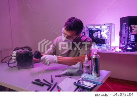 Young man wearing protective mask, gloves glasses performing maintenance on computer power supply, blowing dust away with compressed air at workspace. Young man wearing protective mask, gloves glasses performing maintenance on computer power supply, blowing dust away with compressed air at workspace. 143072104
