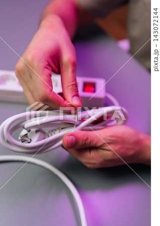Close-up of hands using twist tie to organize white power cord. Power strip with red switch in background. Pink and purple lighting creates modern, tech mood on grey surface. Concept of tidiness. 143072144