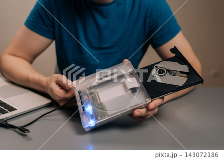 Close-up hand of person holding transparent external optical drive with open disc tray connected to laptop on desk, demonstrating portable dvd cd reading, writing and data transfer for work or home. 143072186