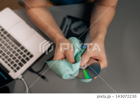 Top view of person cleaning digital disk with microfiber cloth, ensuring data integrity before loading it into an external optical drive connected to a laptop for media playback or storage. 143072230