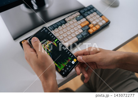 Close-up of trader analyzing real-time market data on smartphone, reviewing stock and crypto charts at desk with keyboard to monitor investments and make informed trading decisions. 143072277