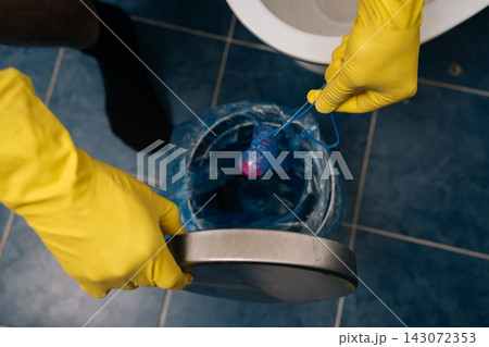 Human hands in yellow protective gloves dropping used toilet bowl cleaner block into lined rubbish bin, highlighting bathroom hygiene, waste disposal and household cleaning routine, close-up. 143072353