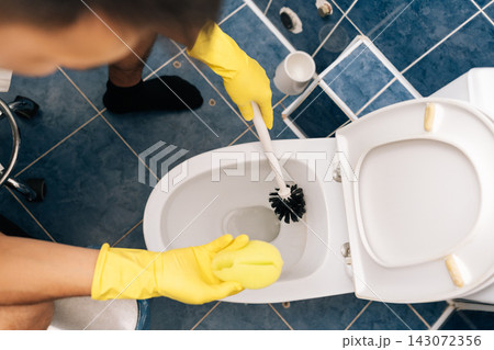 Top view of man wearing yellow protective gloves cleaning toilet bowl with brush, performing essential sanitary maintenance and daily household chores in home bathroom. Concept of household chores. Top view of man wearing yellow protective gloves cleaning toilet bowl with brush, performing essential sanitary maintenance and daily household chores in home bathroom. Concept of household chores. 143072356