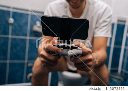 Unrecognizable relaxed young man holding smartphone with gamepad attachment and playing video games sitting on toilet, illustrating mobile gaming addiction, multitasking, privacy in bathroom. Unrecognizable relaxed young man holding smartphone with gamepad attachment and playing video games sitting on toilet, illustrating mobile gaming addiction, multitasking, privacy in bathroom. 143072363