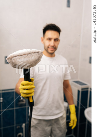 Vertical shot of man in yellow gloves holding active steam cleaner, cleaning surfaces in modern bathroom, focusing on eco-friendly sanitation and maintaining spotless home environment. Vertical shot of man in yellow gloves holding active steam cleaner, cleaning surfaces in modern bathroom, focusing on eco-friendly sanitation and maintaining spotless home environment. 143072365