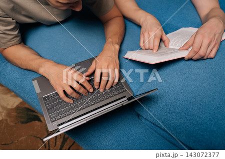 Top view cropped shot of two students studying together on a blue sofa at home, one using a laptop for online learning, the other reading a physical book, both engaged in education. Top view cropped shot of two students studying together on a blue sofa at home, one using a laptop for online learning, the other reading a physical book, both engaged in education. 143072377