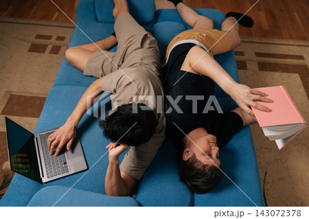 Gay couple relaxing lying on blue sofa, one man engaging with laptop, other engrossed in reading paper book, both creating cozy atmosphere of togetherness during leisure activities at home. Gay couple relaxing lying on blue sofa, one man engaging with laptop, other engrossed in reading paper book, both creating cozy atmosphere of togetherness during leisure activities at home. 143072378