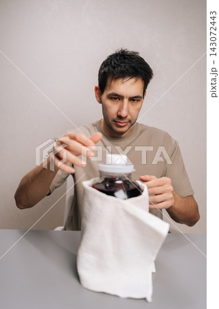 Vertical portrait of young adult man opening homemade compote jar, holding large glass container with white lid, preparing refreshing fruit beverage in kitchen, emphasizing preservation. 143072443