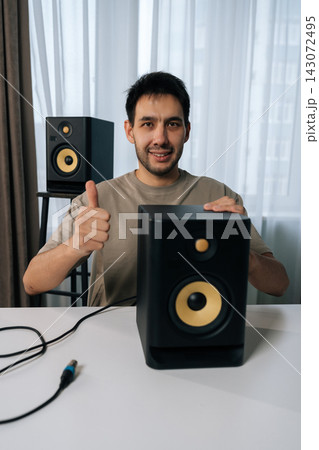 Vertical shot of man smiling looking camera and giving thumbs up gesture, connecting studio monitor speaker for home recording or music production setup, expressing satisfaction with audio equipment. 143072495