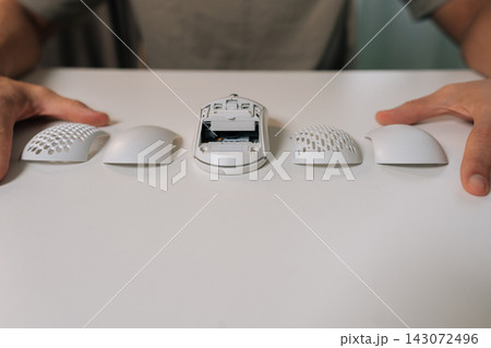 Close-up of person hands on white desk showing disassembled ergonomic wireless computer mouse with core unit and four interchangeable shells, highlighting modular, customizable lightweight design. 143072496