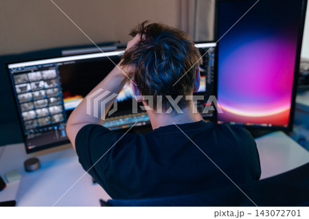 Man feeling stressed and frustrated, gripping head while working on computer with multiple screens in dark home office, experiencing burnout and overwhelming deadlines late at night. 143072701