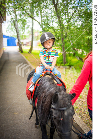 Beautiful little girl two years old riding pony horse in big safety jockey helmet posing outdoors on countryside Beautiful little girl two years old riding pony horse in big safety jockey helmet posing outdoors on countryside 143072938