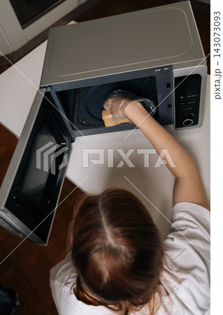High-angle view of housewife in clear disposable gloves scrubbing inside of microwave with yellow sponge, close-up of routine kitchen cleaning and appliance hygiene maintenance. 143073093