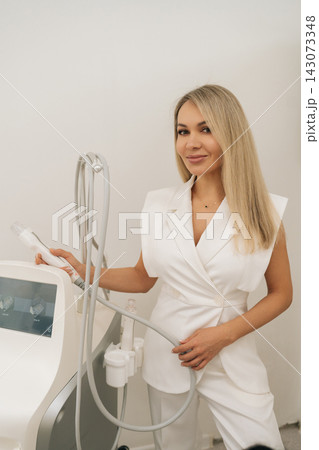 Vertical portrait of blonde esthetician in white coat setting up modern fractional radiofrequency microneedling device, adjusting parameters before facial rejuvenation therapy, looking at camera. Vertical portrait of blonde esthetician in white coat setting up modern fractional radiofrequency microneedling device, adjusting parameters before facial rejuvenation therapy, looking at camera. 143073348