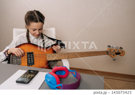 Portrait of cute preteen girl with pigtails focused on playing bass guitar, following online lesson on smartphone, developing new musical skills and a creative hobby at home. 143073425