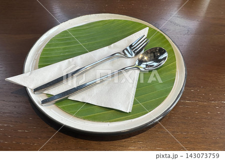 top view cutlery placed on a triangular folded paper napkin, set on a banana leaf atop a white ceramic plate, resting on a wooden floor background, object, decor, copy space 143073759