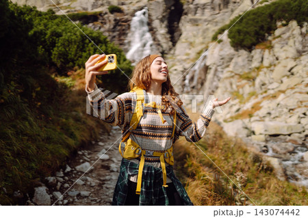 A woman with a yellow backpack smiles as she takes a picture with her phone near a waterfall. 143074442