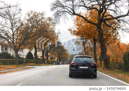 Back scenic view black family car driving wet countryside road through bright autumnal golden red orange nature foliage forest trees. Scenery fall season highway vehicle. Drive auto safety 143074669
