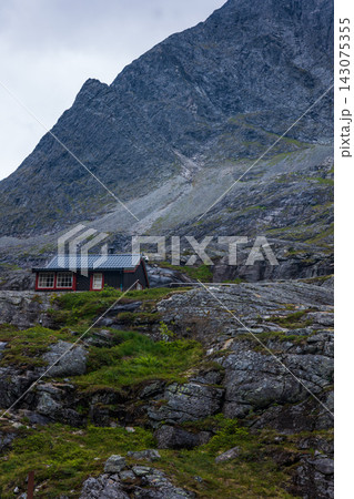 Red wooden cottage beneath mountain slope in Norwegian valley, moss-covered rocks Red wooden cottage beneath mountain slope in Norwegian valley, moss-covered rocks 143075355