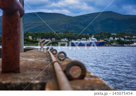 Mooring post and chain on pier with cargo ship, Norwegian fjord settlement and mountains 143075454