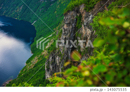 Aerial view of steep rocky cliff with dense forest vegetation above deep blue fjord water, Norway Aerial view of steep rocky cliff with dense forest vegetation above deep blue fjord water, Norway 143075535