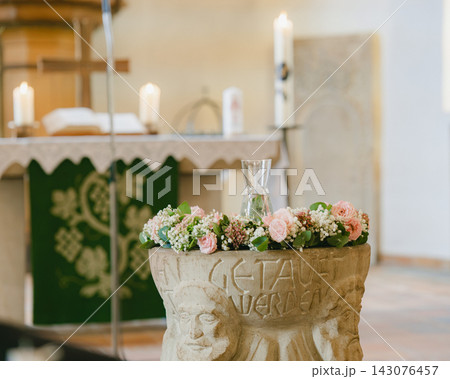 Ancient stone baptismal font decorated with fresh floral wreath of roses and gypsophila inside light church interior. Preparation for infant baptism sacrament and christening ceremony Ancient stone baptismal font decorated with fresh floral wreath of roses and gypsophila inside light church interior. Preparation for infant baptism sacrament and christening ceremony 143076457