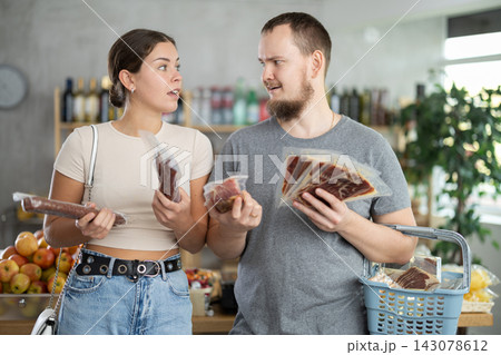 Satisfied male and female customers with dried sausage and jamon in hands shopping together in grocery store Satisfied male and female customers with dried sausage and jamon in hands shopping together in grocery store 143078612