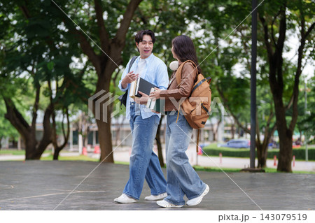 Two Asian college students walking together across green university campus carrying textbooks 143079519
