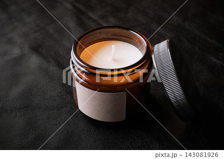 Scented candle in an amber glass jar with blank label, lid resting on black background 143081926