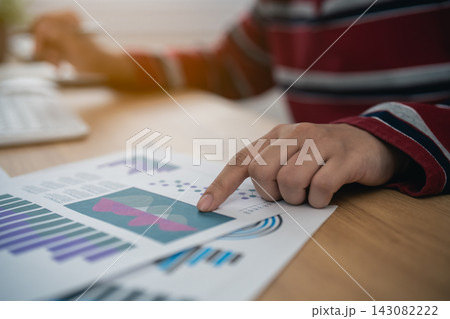 Person analyzing business chart report data with finger pointing on graph paper on wooden desk in office workspace Person analyzing business chart report data with finger pointing on graph paper on wooden desk in office workspace 143082222
