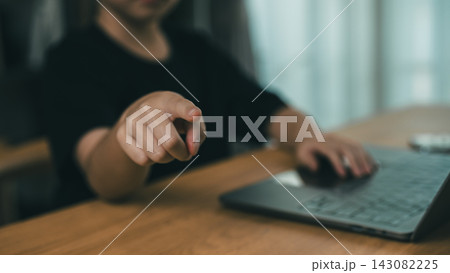 Child pointing finger towards camera sitting at wooden desk with laptop computer in blurred background indoors Child pointing finger towards camera sitting at wooden desk with laptop computer in blurred background indoors 143082225