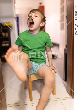 Child sits on chair with one foot up while yawning and holding a snack in his mouth in indoor setting during daytime 143086849