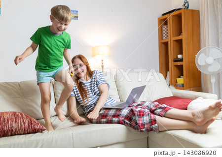 Boy jumps on couch while mother works on laptop in living room during afternoon 143086920