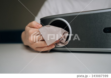 Person hand wiping projector lens with specialized microfiber cloth. Ensuring proper care and maintenance of the home cinema equipment for clear viewing and extending lifespan, close-up, cropped shot. 143087399