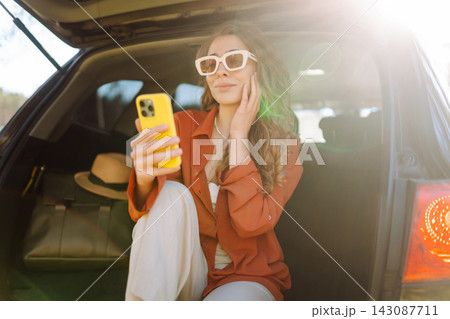 Beautiful woman sits in the trunk of a car in the forest and uses a smartphone. Traveling by car Beautiful woman sits in the trunk of a car in the forest and uses a smartphone. Traveling by car 143087711