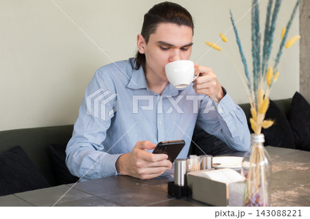 A young man is sitting at a table in a cafe. He looks at his phone and holds a cup 143088221