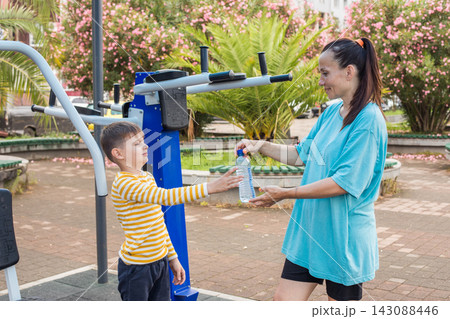 Smiling child and woman holding water from a plastic bottle during workouts in outdoor gym park 143088446