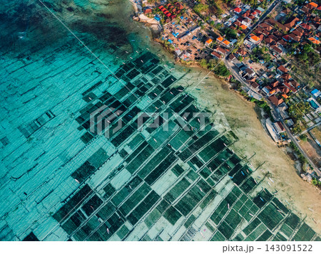 Drone view of turquoise coastal aquaculture farms beside a tropical seaside village 143091522