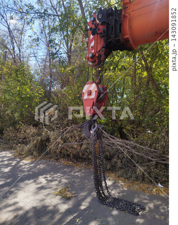 Close-up of a bright orange mobile crane hook block with heavy steel chains hanging down to the asphalt road in an outdoor location surrounded by trees under a sunny sky. Close-up of a bright orange mobile crane hook block with heavy steel chains hanging down to the asphalt road in an outdoor location surrounded by trees under a sunny sky. 143091852